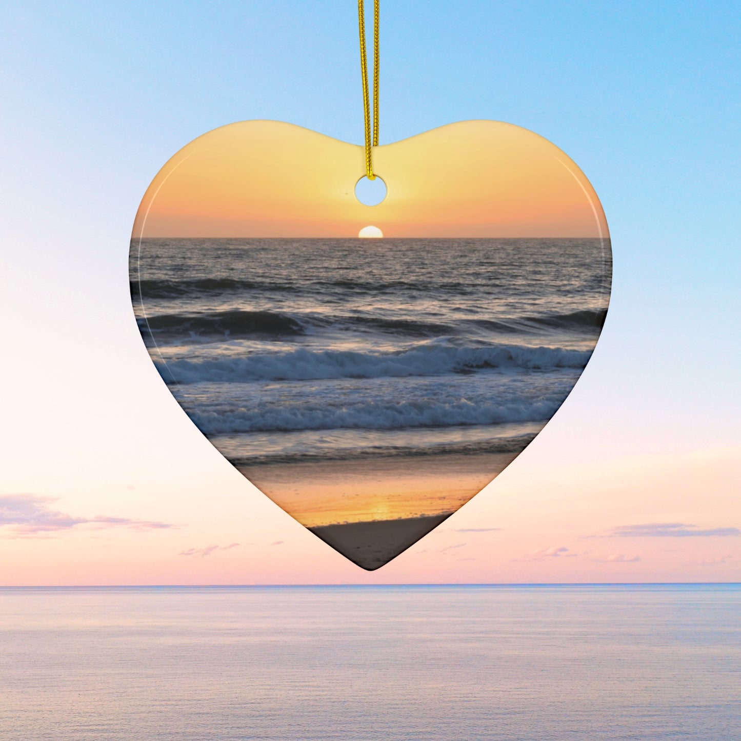 Heart-shaped ceramic Christmas ornament featuring a photograph of a vibrant amber and gold sunset over the Pacific Ocean with waves rolling toward the shore.