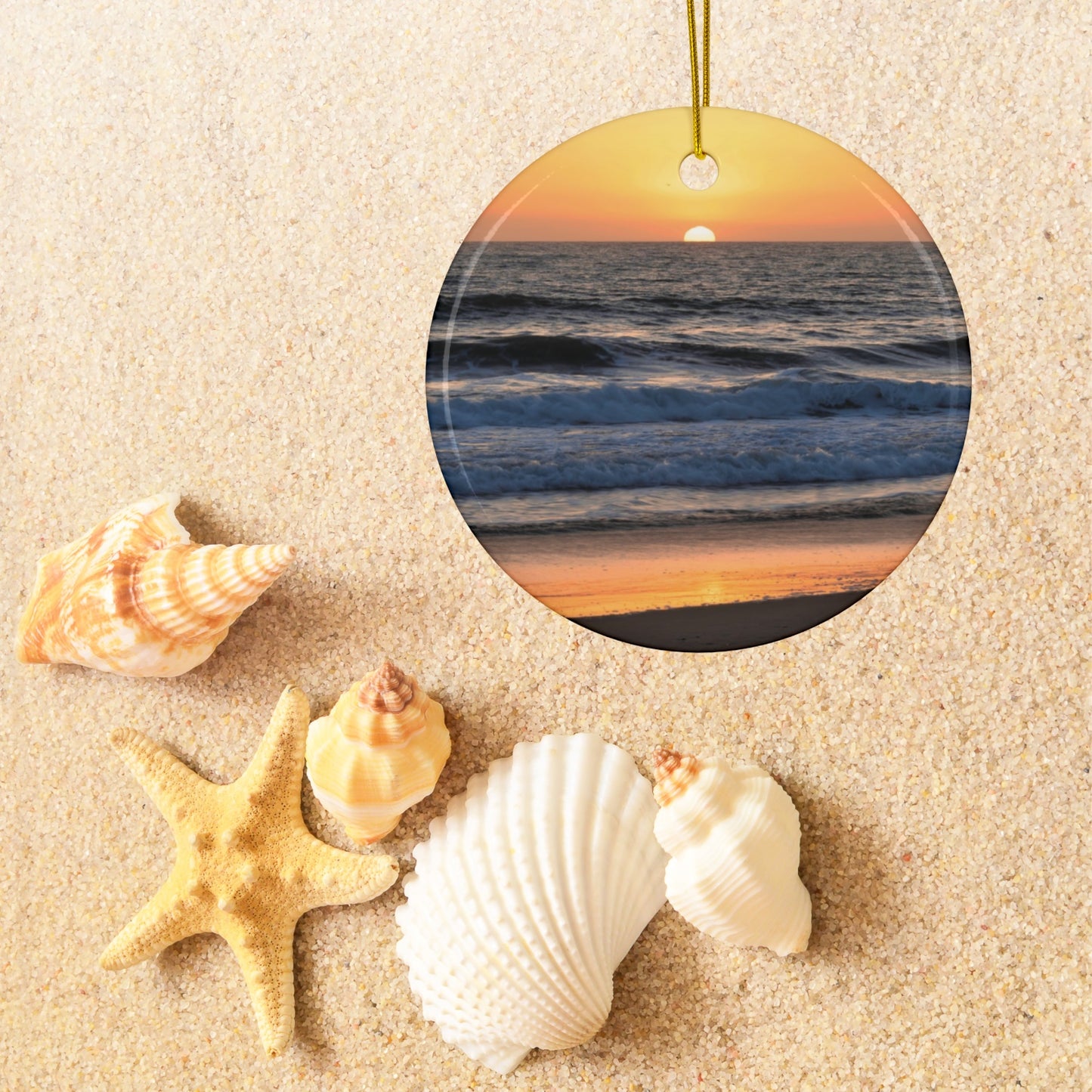 Ceramic Christmas ornament featuring a photograph of a vibrant amber and gold sunset over the Pacific Ocean with waves rolling toward the shore on sand with shells and a starfish