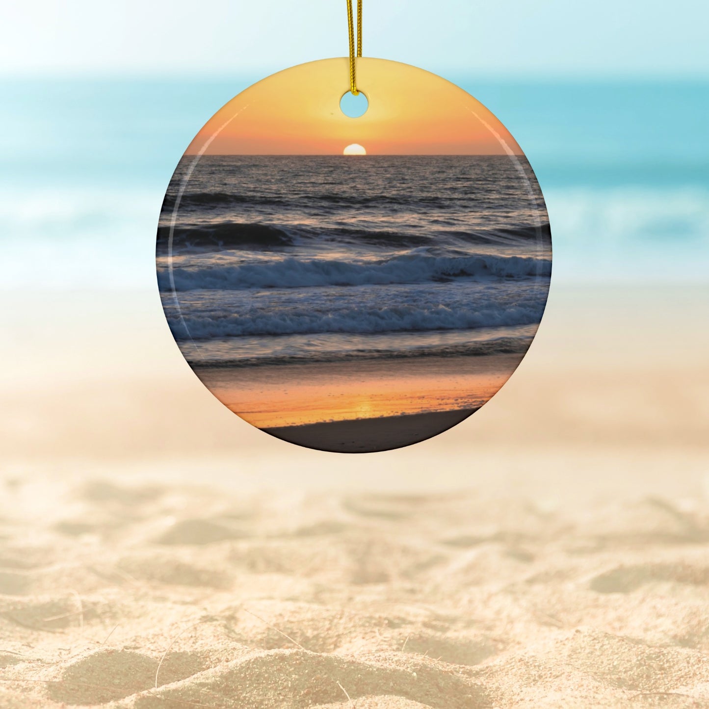 Ceramic Christmas ornament featuring a photograph of a vibrant amber and gold sunset over the Pacific Ocean with waves rolling toward the shore.