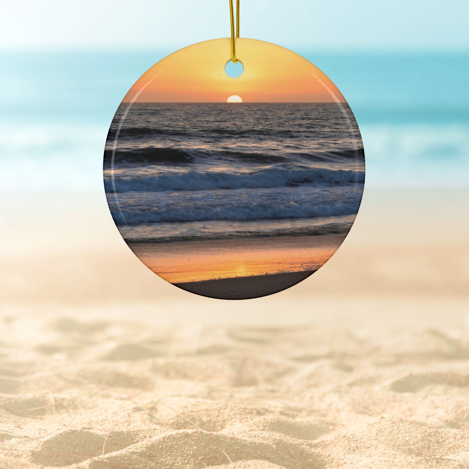 Ceramic Christmas ornament featuring a photograph of a vibrant amber and gold sunset over the Pacific Ocean with waves rolling toward the shore.