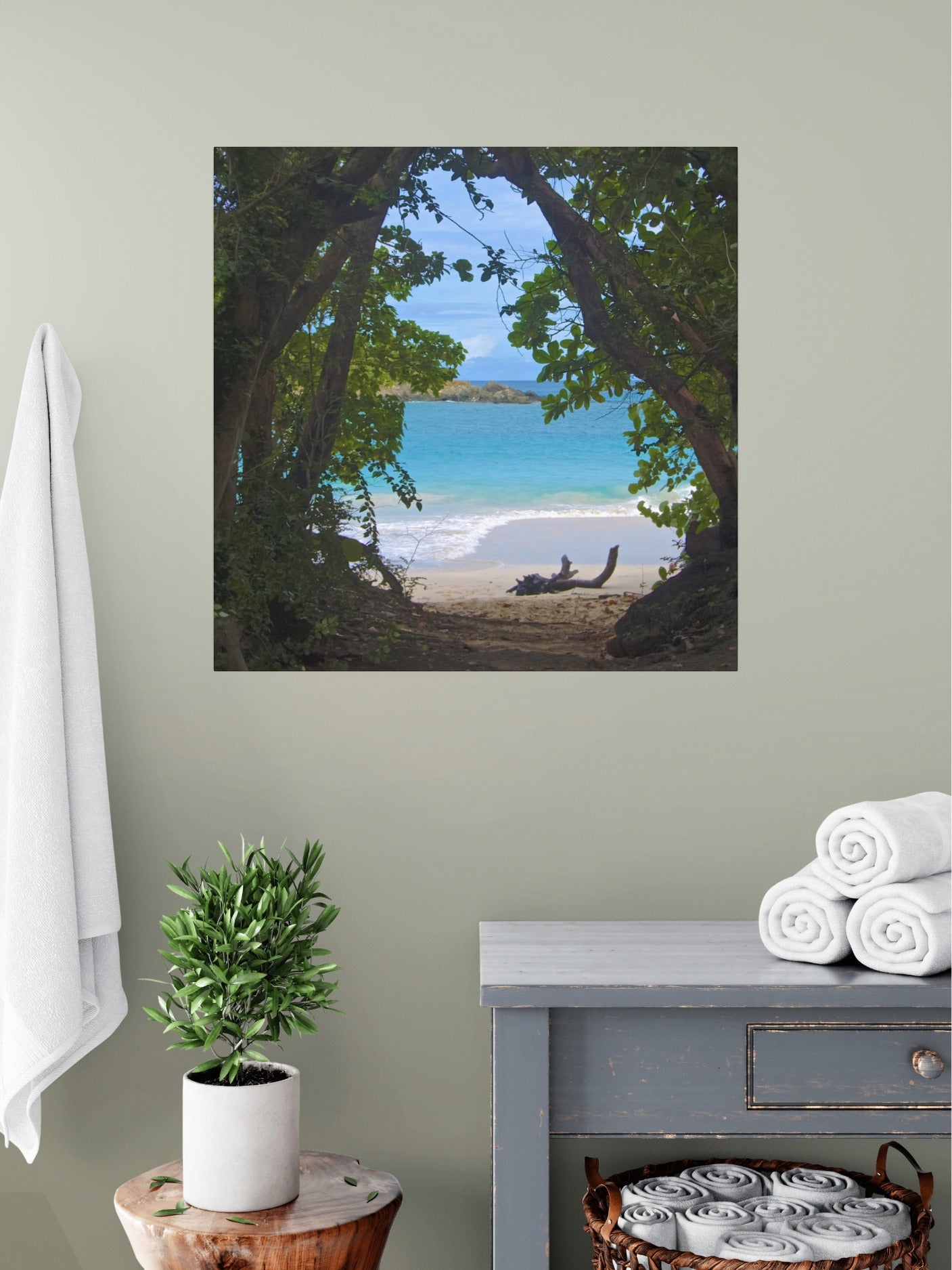 Tropical beach view through trees Hawaiian island canvas print on a wall above a dresser with towels and a plant.