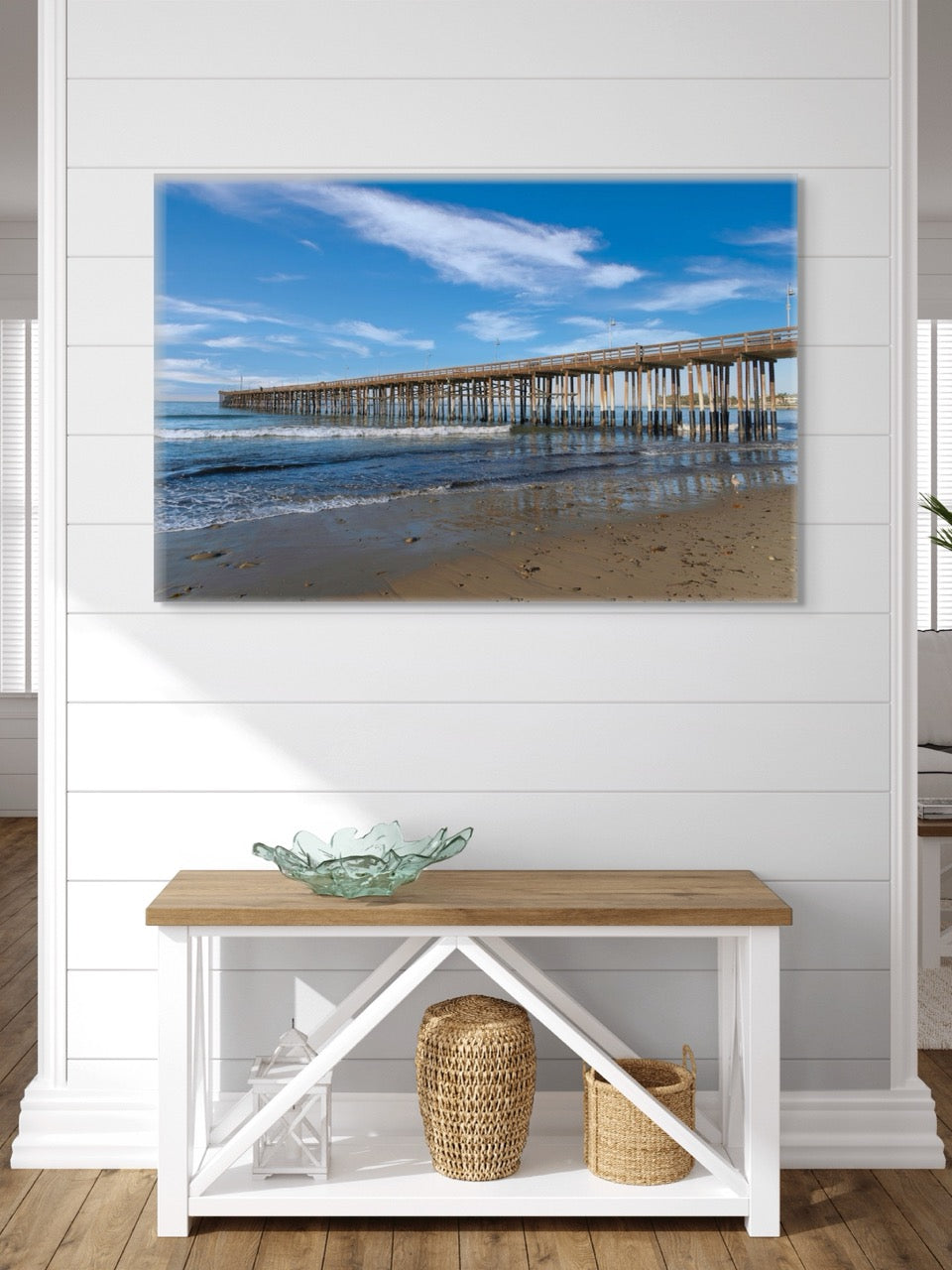 Large canvas print of Ventura Pier in California on a wall above a wooden bench with a glass bowl and baskets.