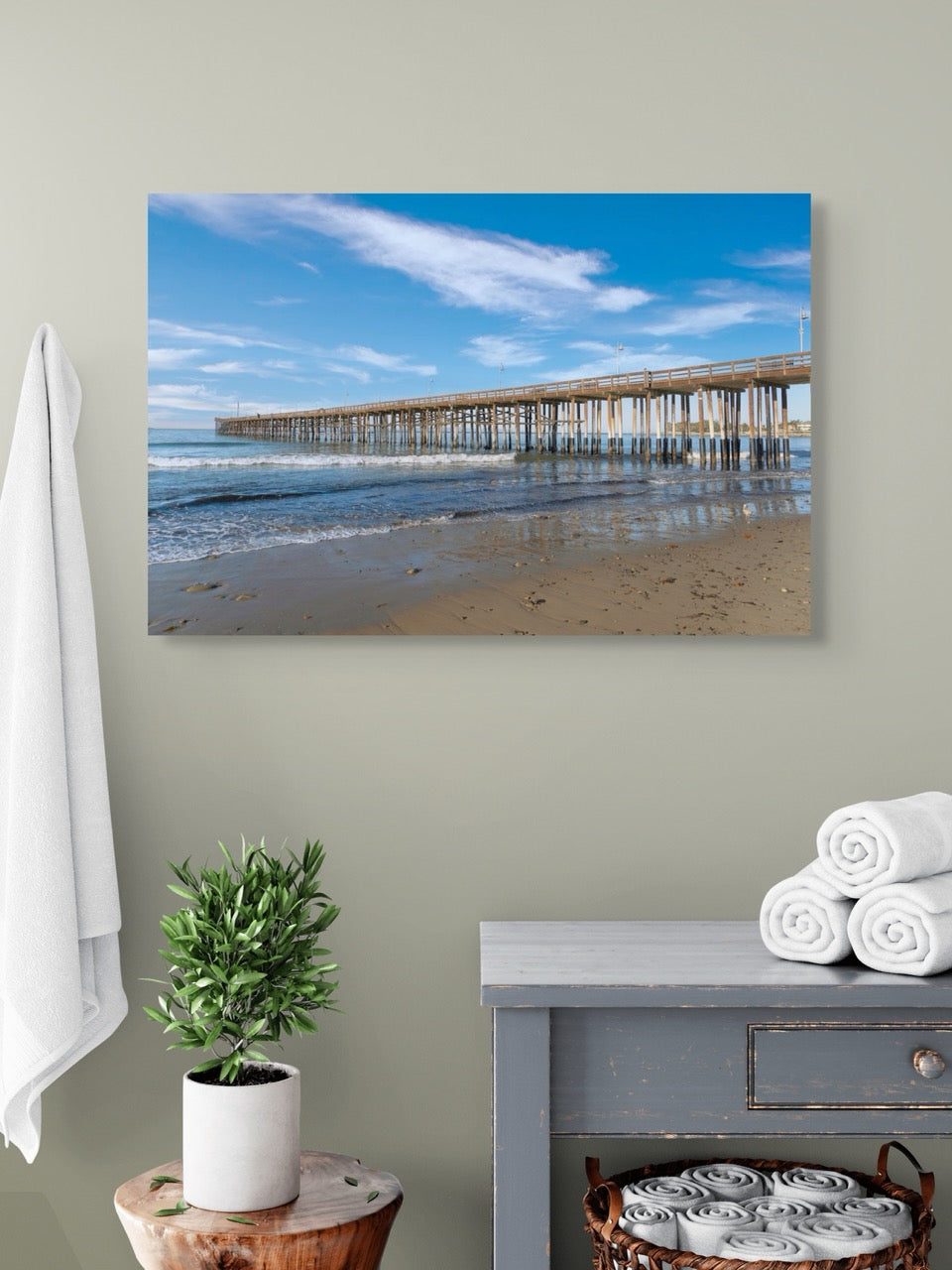 A canvas print of Ventura Pier in California on wall above a table with a plant and folded towels.