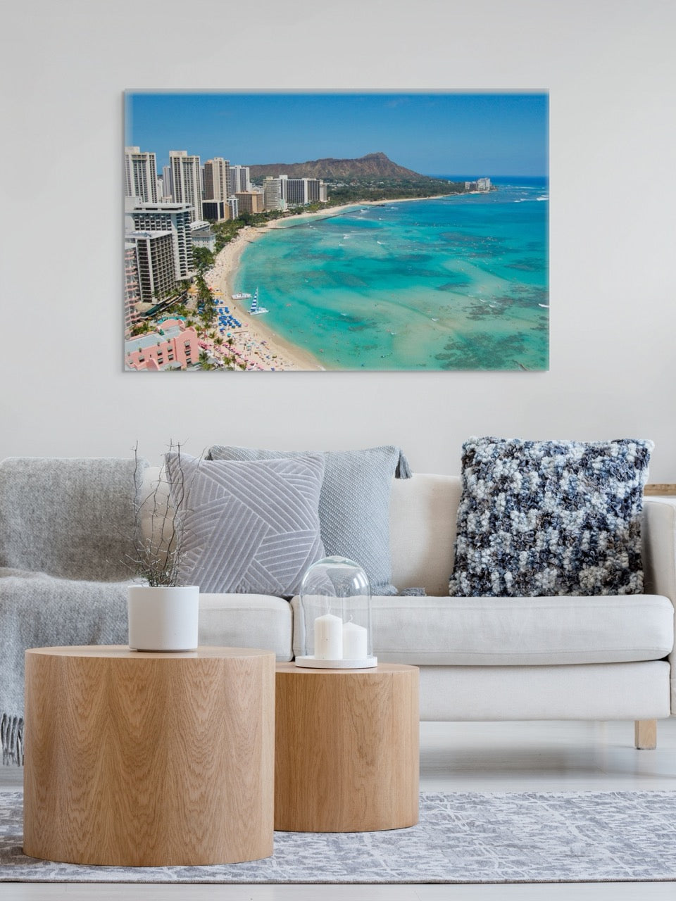 Living room with a large canvas print of an overhead view of the beach and hotels on Waikiki Beach in Hawaii with Diamond Head in the background, a sofa with decorative pillows, and wooden side tables.