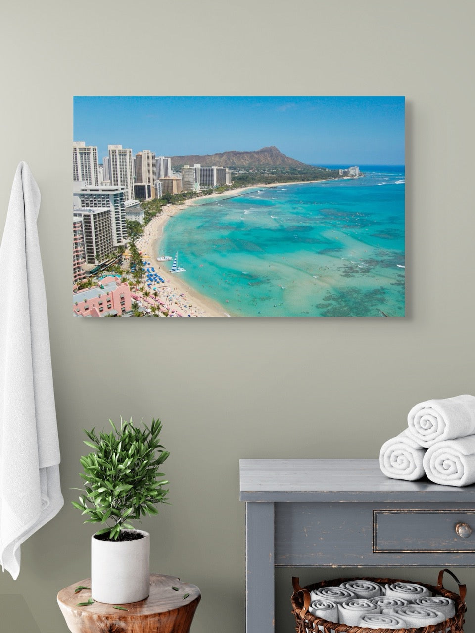 canvas print of an overhead view of the beach and hotels on Waikiki Beach in Hawaii with Diamond Head in the background, hanging on a wall above a table with folded towels.