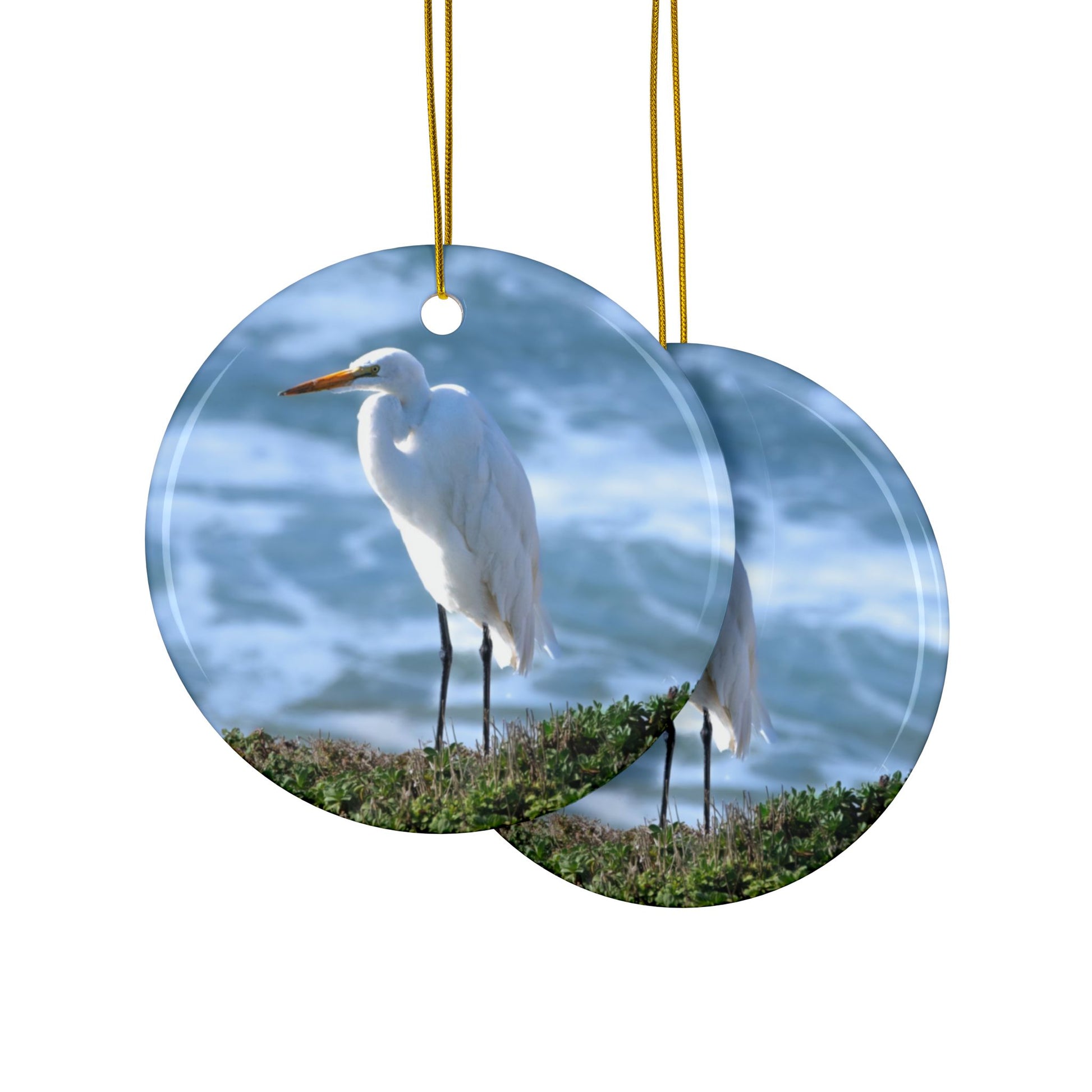 Front and back side of a ceramic holiday ornament featuring a  photograph of a White Egret on a cliff overlooking blue ocean waves with sea spray