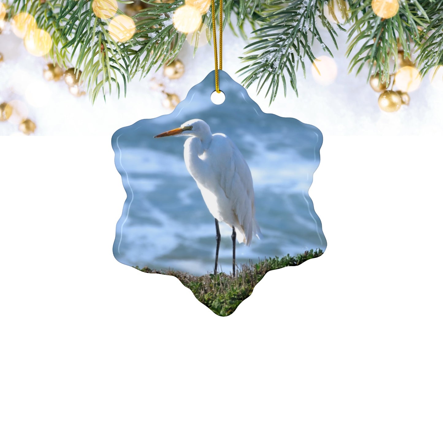 Ceramic holiday ornament featuring a  photograph of a White Egret on a cliff overlooking blue ocean waves with sea spray, hanging from a branch with lights.