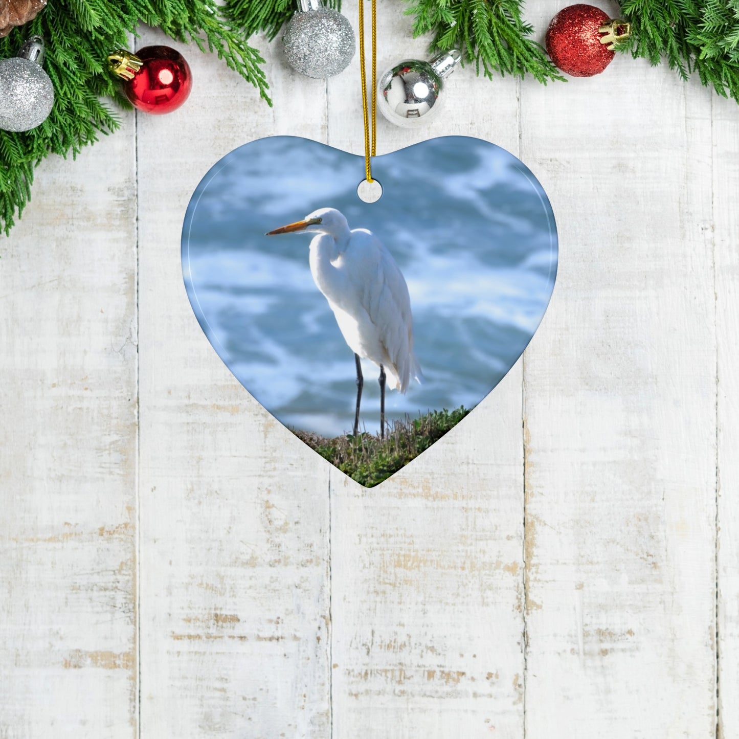 Heart-shaped ceramic holiday ornament featuring a  photograph of a White Egret on a cliff overlooking blue ocean waves with sea spray, hanging against a wooden background with Christmas decorations.