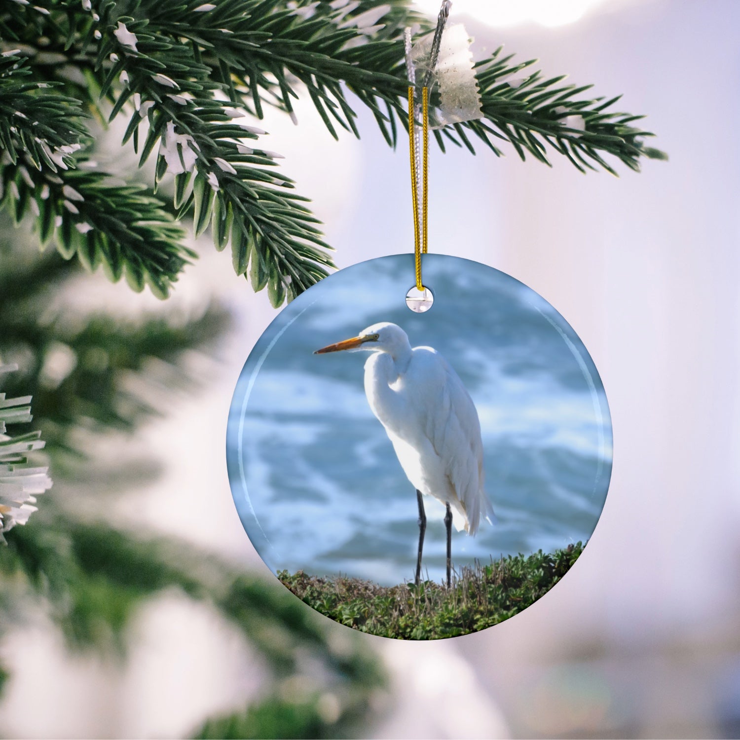 Ceramic holiday ornament featuring a  photograph of a White Egret on a cliff overlooking blue ocean waves with sea spray hanging on a tree branch.