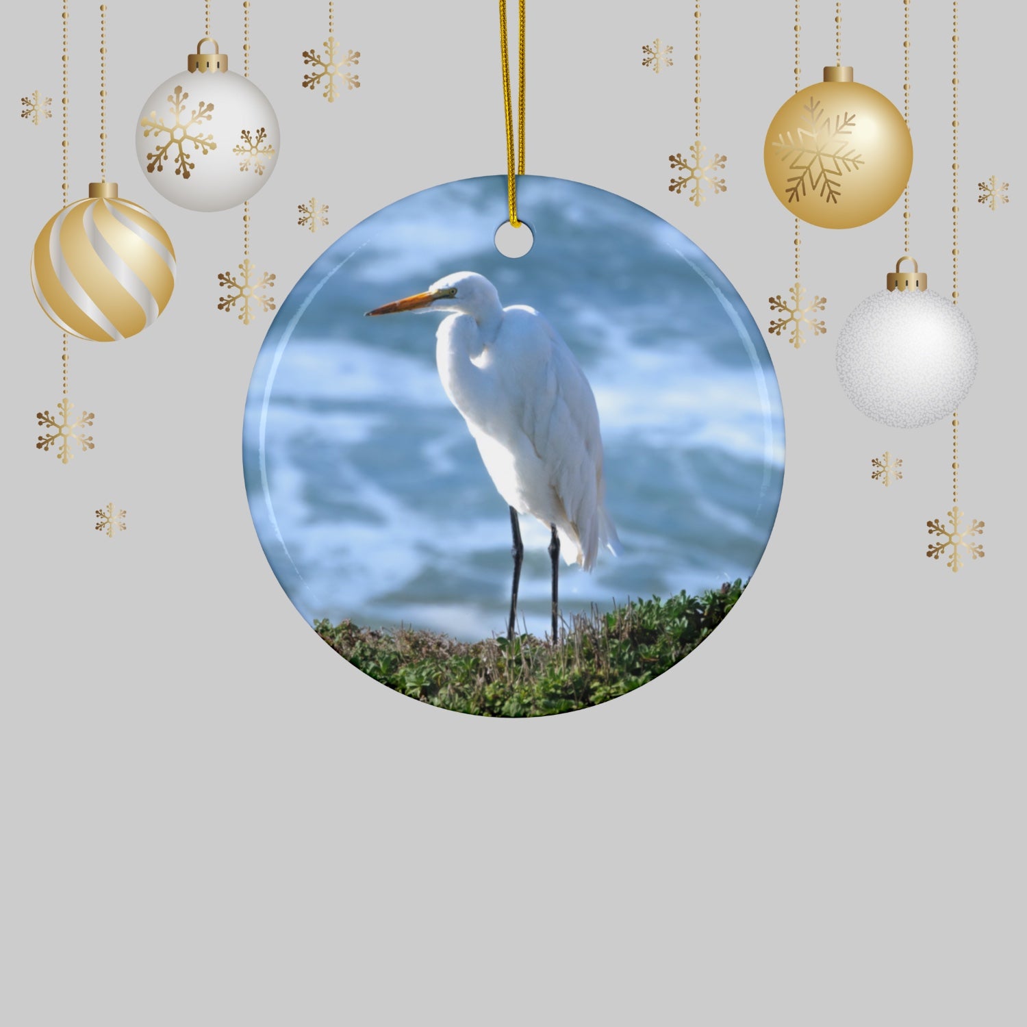 Ceramic holiday ornament featuring a  photograph of a White Egret on a cliff overlooking blue ocean waves with sea spray and Christmas ornaments on a light gray background