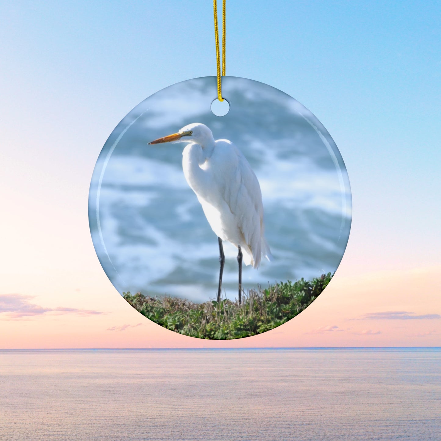 Ceramic holiday ornament featuring a  photograph of a White Egret on a cliff overlooking blue ocean waves with sea spray