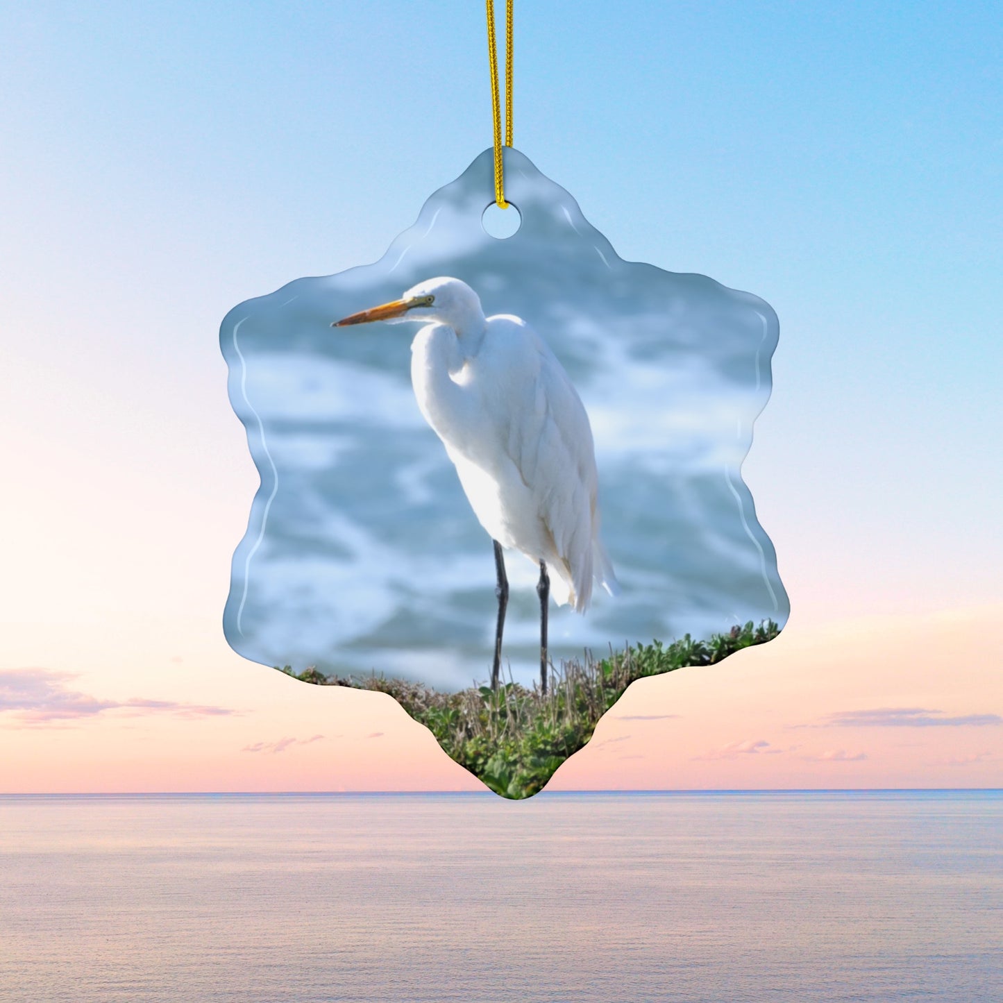Ceramic holiday ornament featuring a  photograph of a White Egret on a cliff overlooking blue ocean waves with sea spray