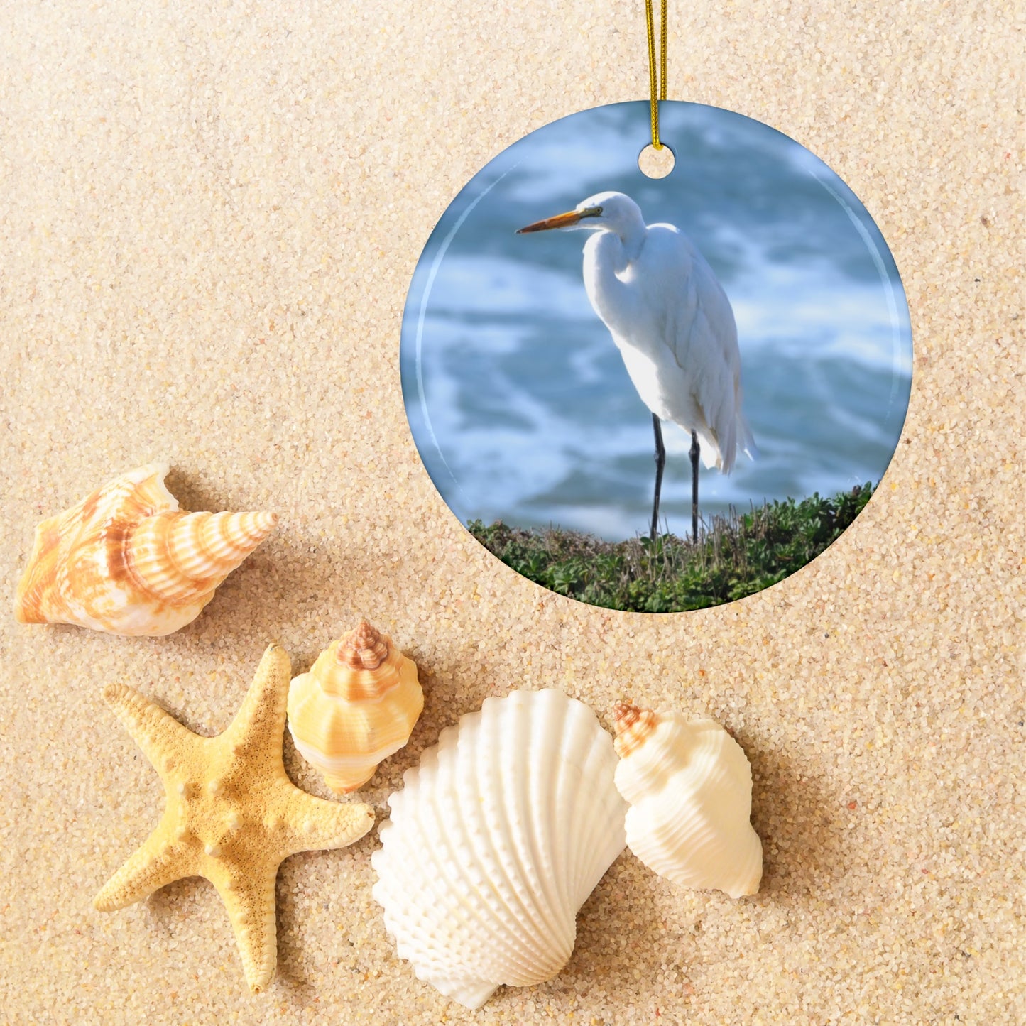Ceramic holiday ornament featuring a  photograph of a White Egret on a cliff overlooking blue ocean waves with sea spray surrounded by shells and a starfish.