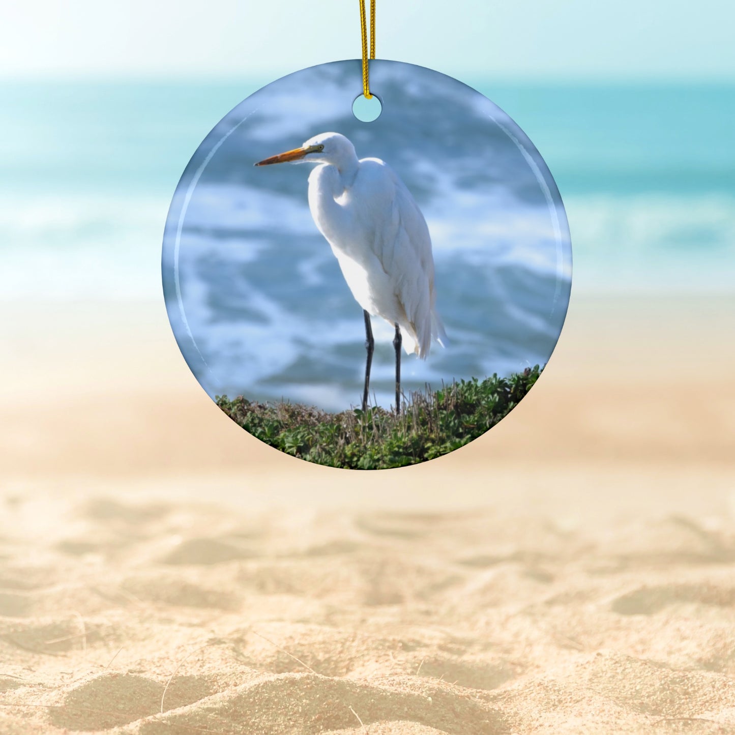 Ceramic holiday ornament featuring a  photograph of a White Egret on a cliff overlooking blue ocean waves with sea spray