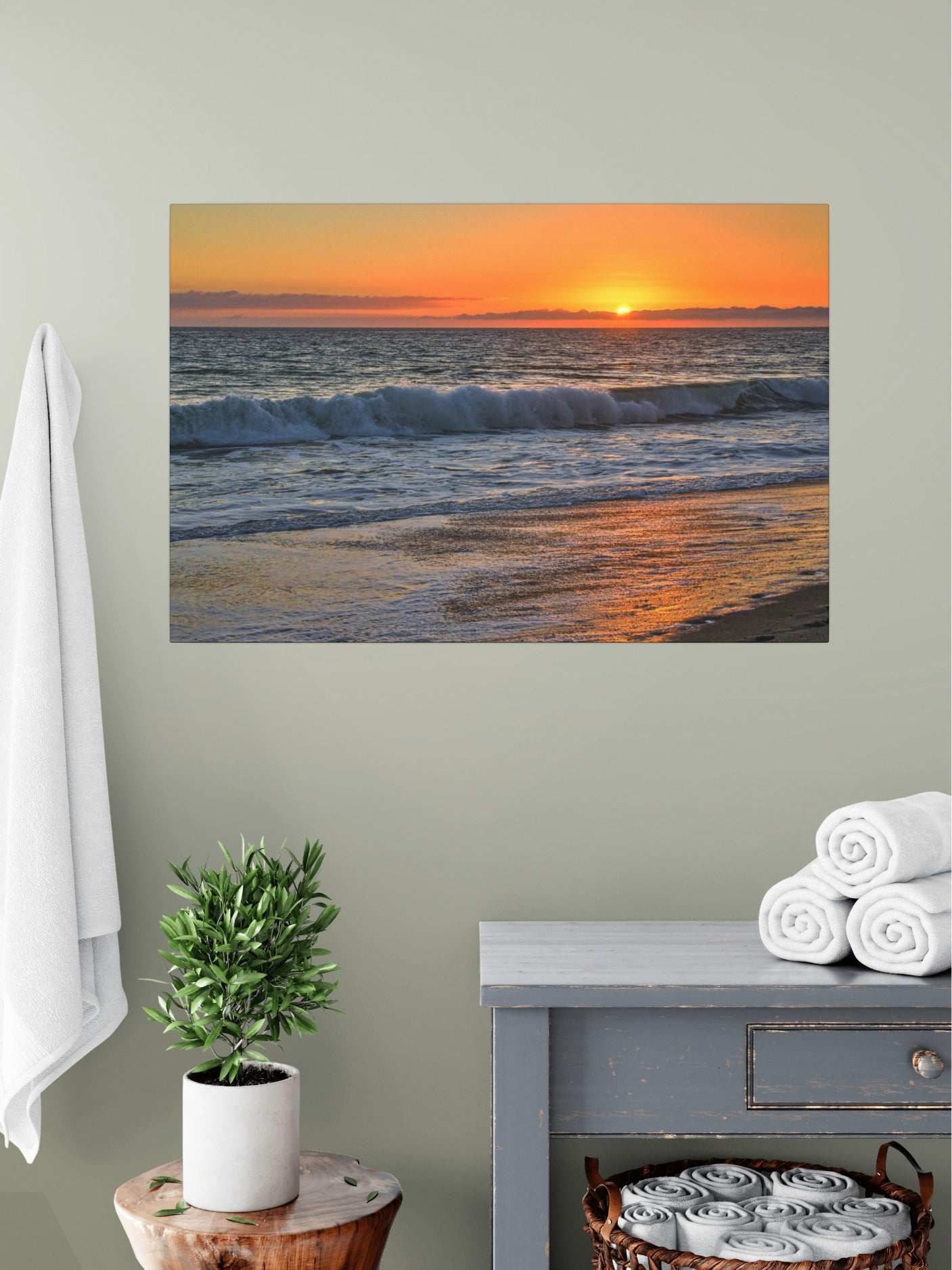 Canvas print of a Zuma Beach sunset in Malibu California with golden sky and reflective sand on wall above a table with a plant and folded towels.